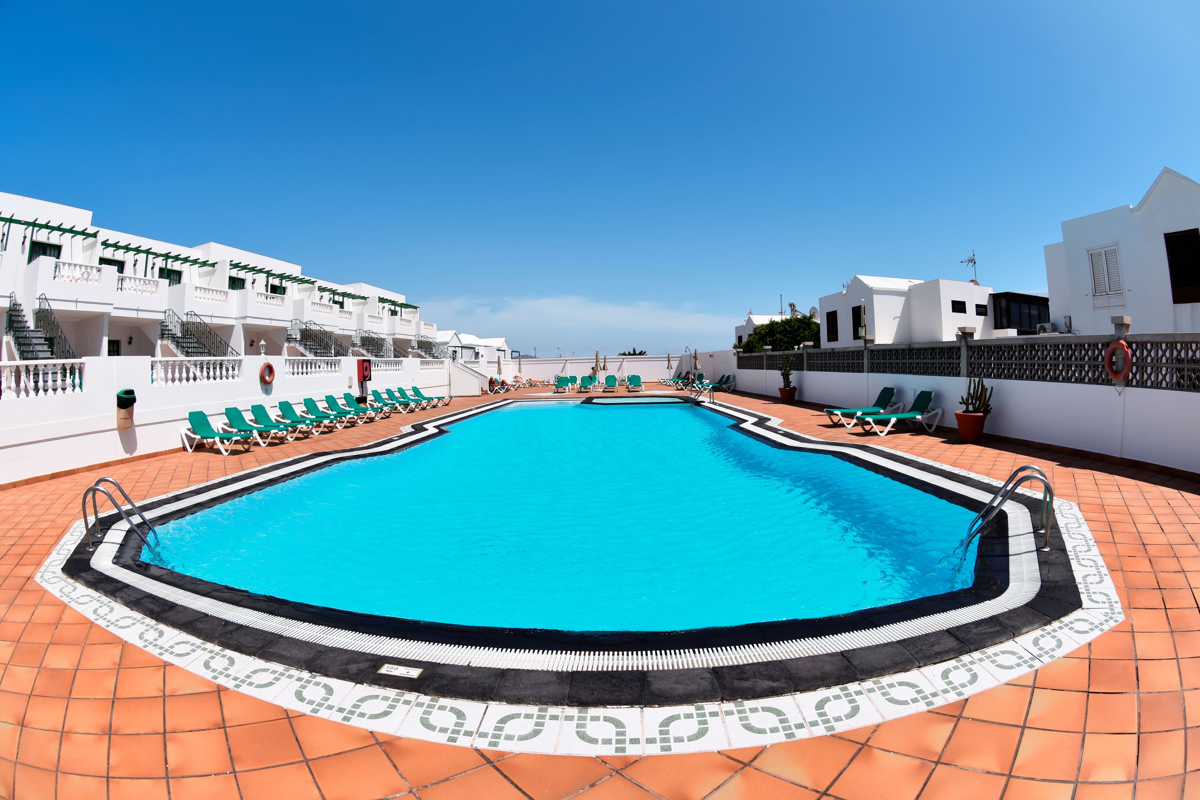 Une piscine dotée d’une terrasse carrelée, entourée de chaises longues et bordée d’un bâtiment blanc avec des balcons.