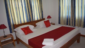 Un lit double avec une couvre-lit rouge et des oreillers, une tête de lit en bois, deux tables de chevet avec des lampes et des rideaux à motifs bleus et jaunes.