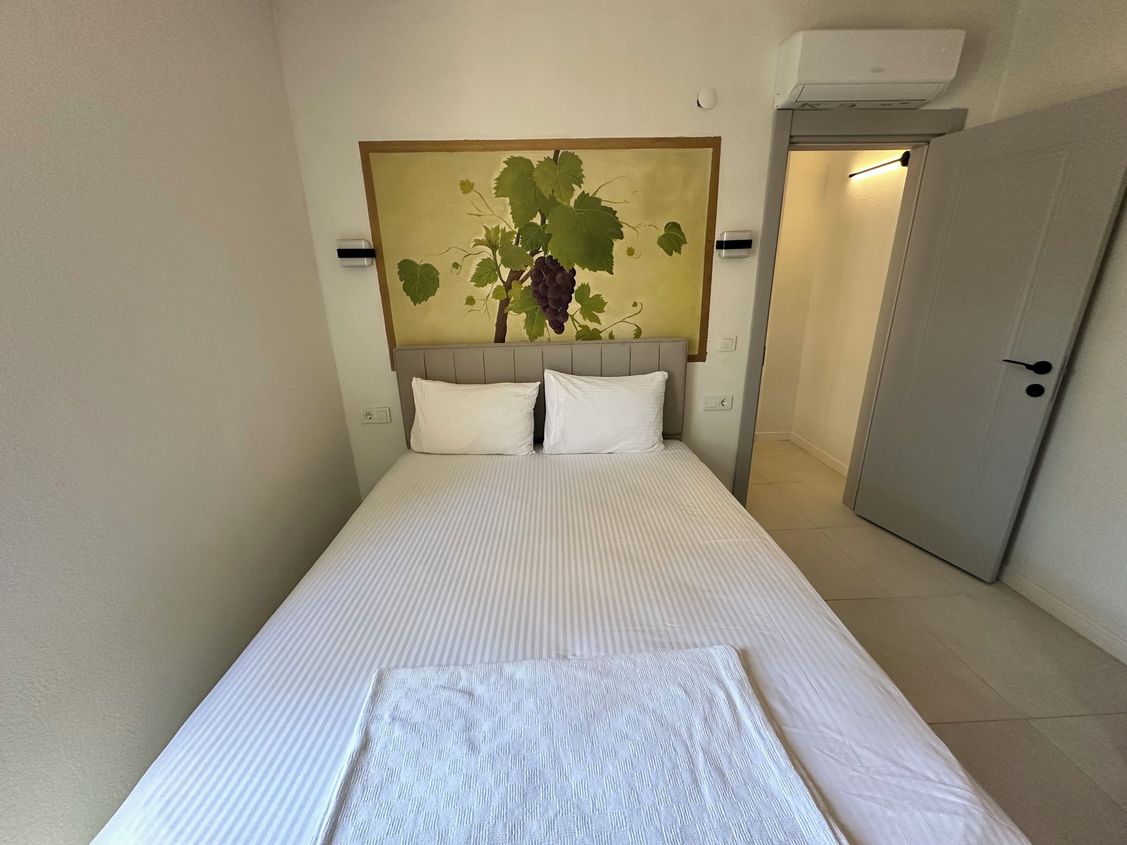 A neatly made bed with a striped bedspread, a headboard with a grapevine mural, and a closed door to the right.