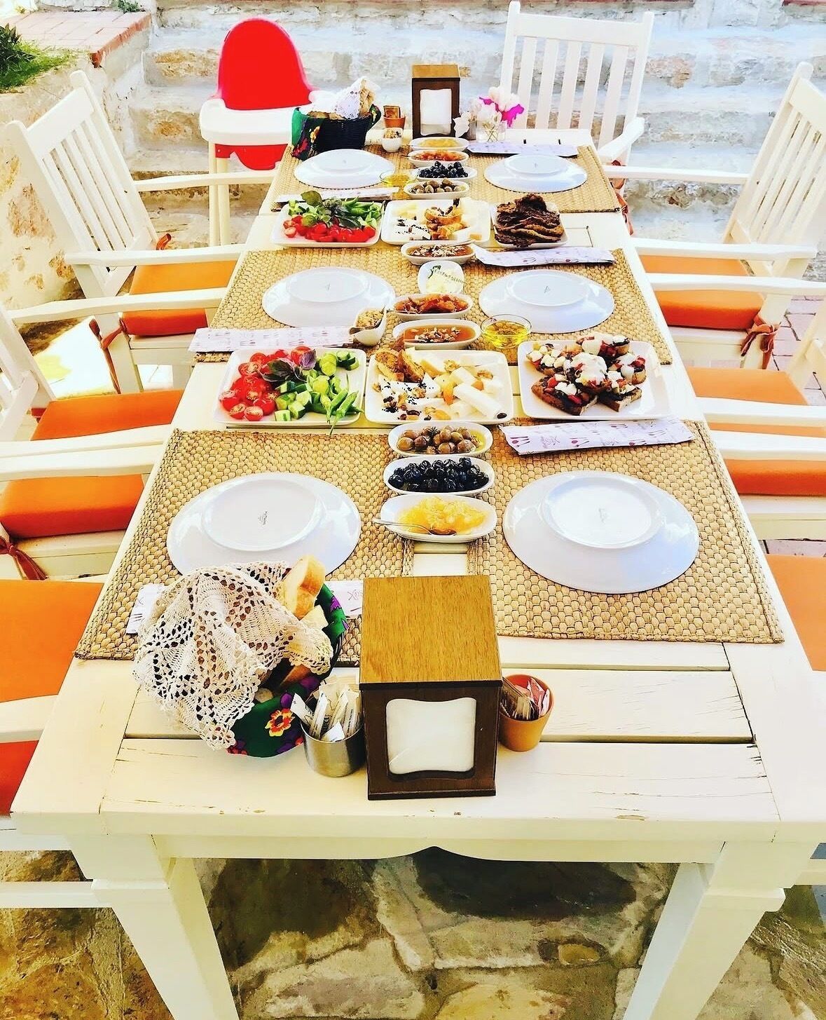 Una mesa preparada para una comida con varios platos, sillas blancas con cojines anaranjados y un camino de mesa de mimbre.
