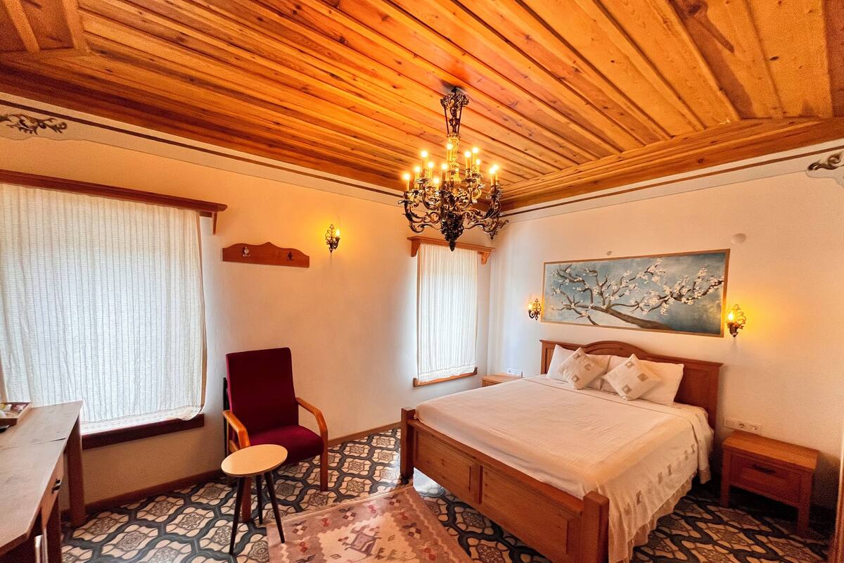 A room with a wooden ceiling, a bed, a chair, a desk, and a painting on the wall.