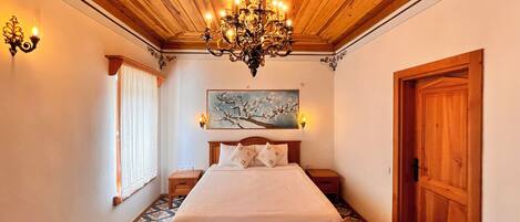 A bedroom with a wooden ceiling, a chandelier, a bed with white bedding, a nightstand, a wall-mounted painting, and a patterned floor.