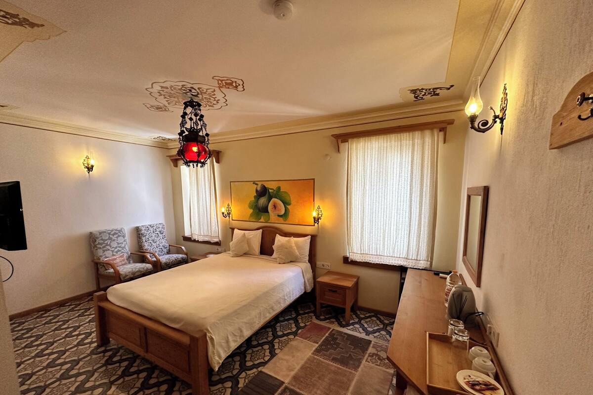 A hotel room with a bed, two chairs, a bedside table, a nightstand, a lamp, a painting, and a mirror.