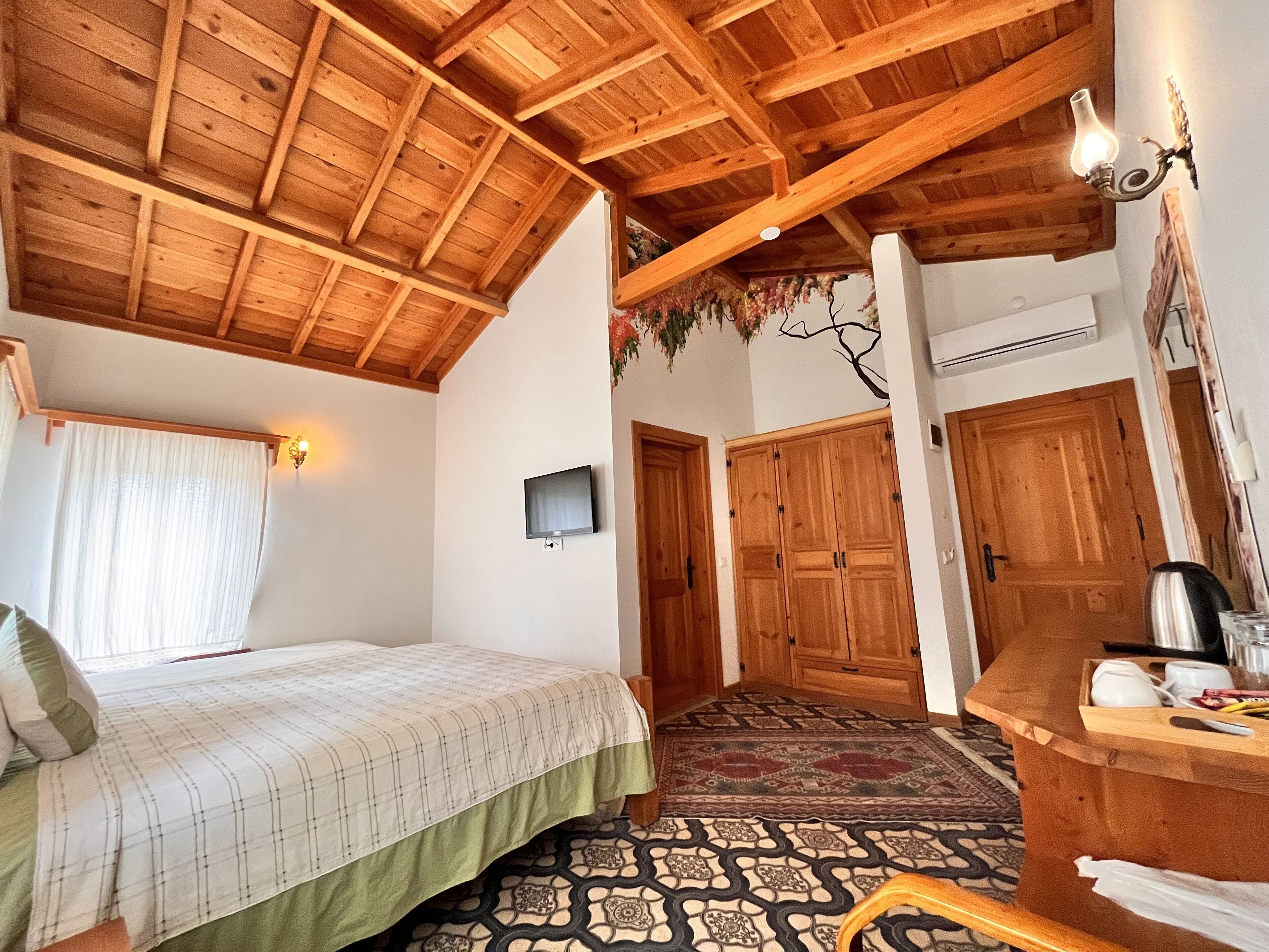 Un dormitorio con techo de madera, una cama con colcha verde, un televisor montado en la pared y un escritorio de madera con hervidor.