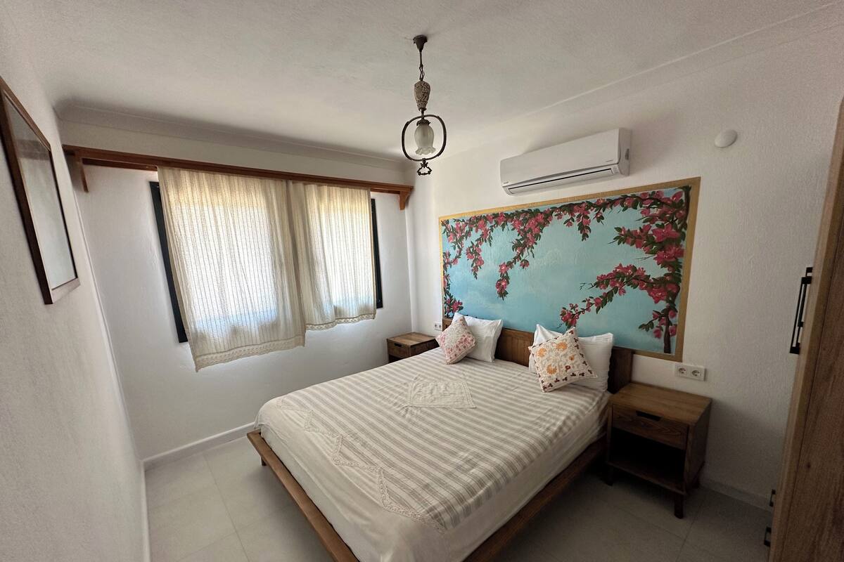 A bedroom with a bed, two nightstands, a ceiling fan, an air conditioning unit, a window with curtains, and a floral wall mural.