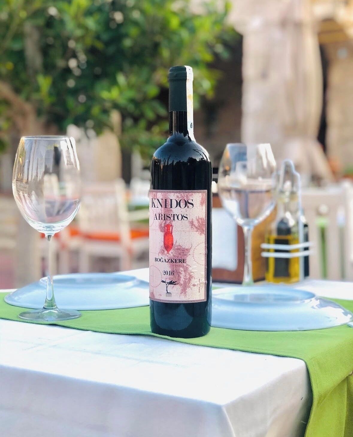 Una botella de vino Knidos Aristos sobre una mesa con mantel verde, una copa de vino y otra botella al fondo.