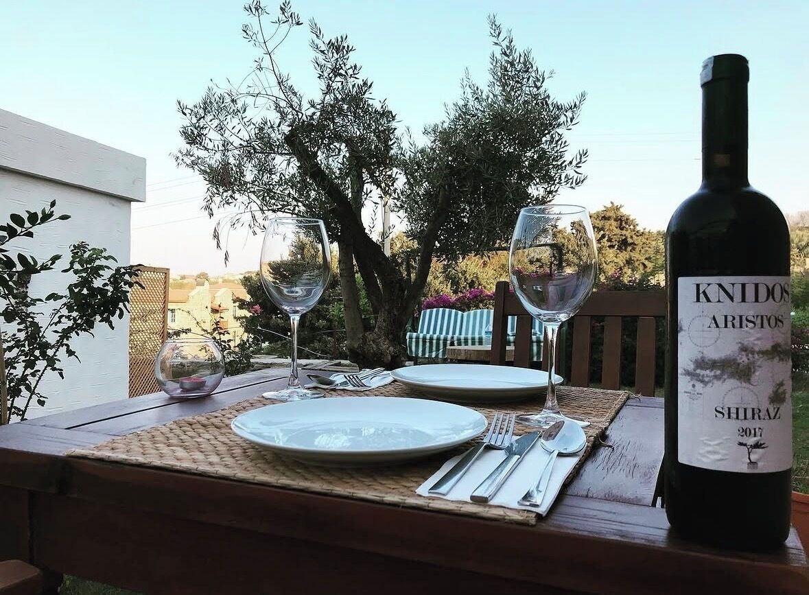 Une table dressée à l’extérieur, avec une bouteille de vin KNIDOS ARISTOS Shiraz 2017, deux verres à vin, des assiettes, des couverts et un set de table en fibres tressées.