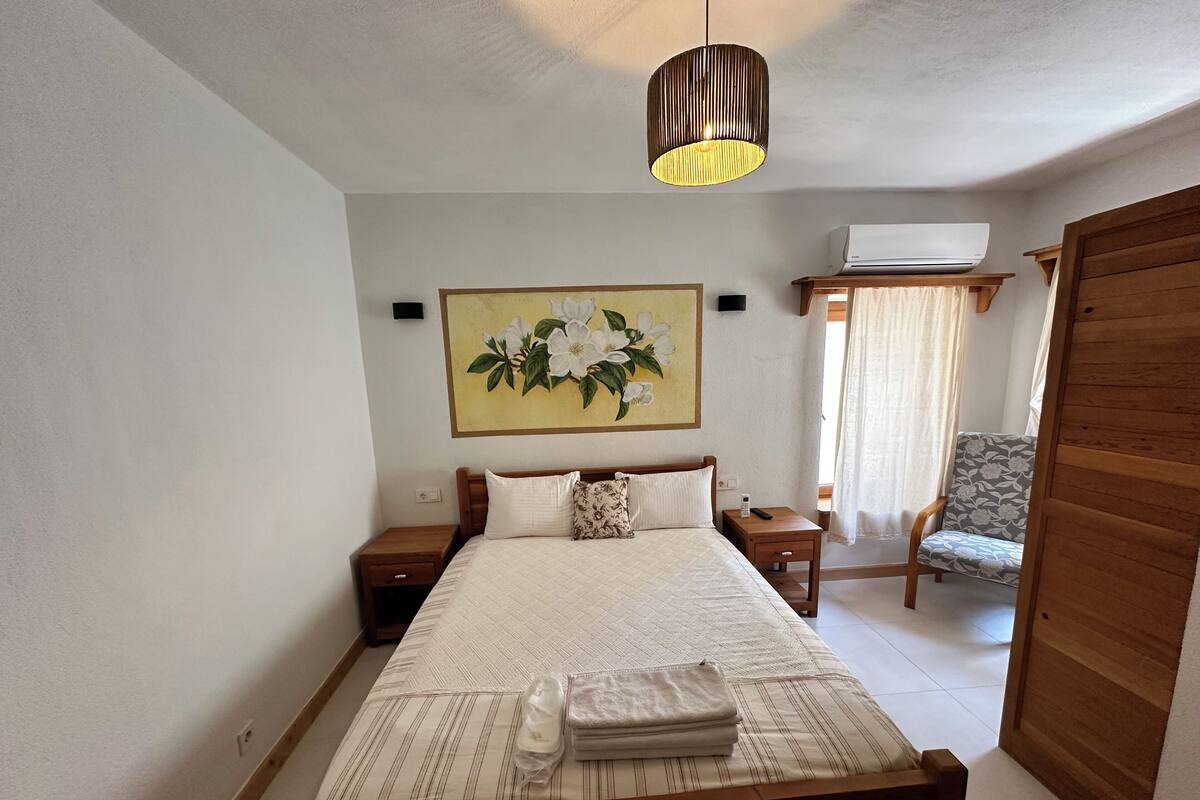 A bedroom with a bed, a nightstand, a wall-mounted air conditioner, a chair, and a painting.