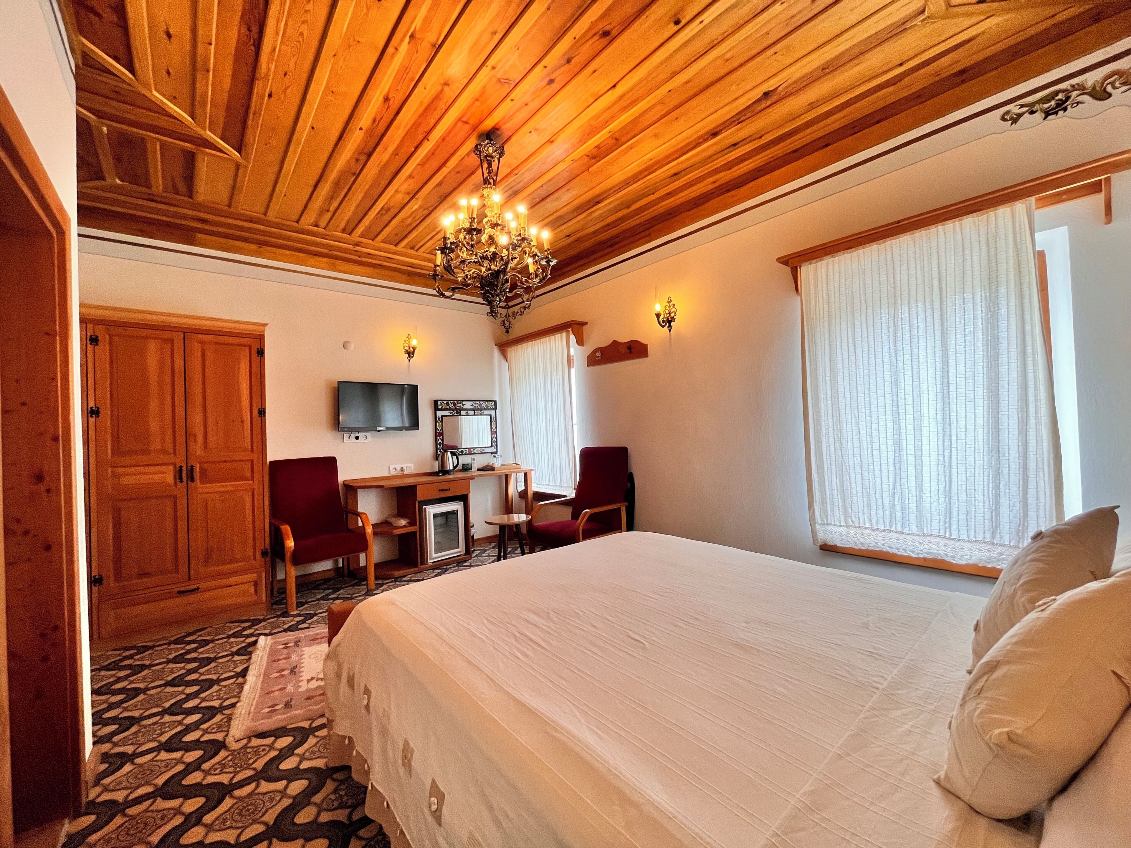 A hotel room with a wooden ceiling, a bed, a desk with chairs, a television, and a window with curtains.
