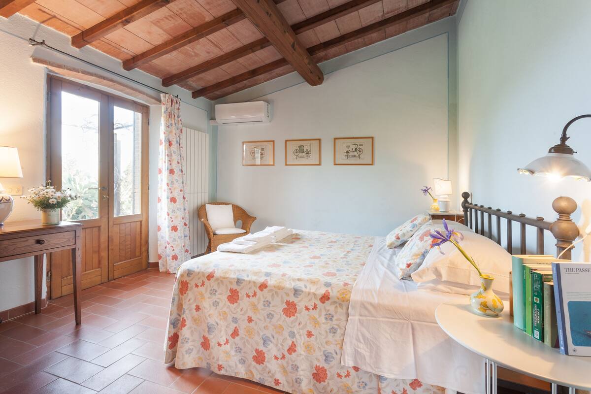 Een slaapkamer met een bed, nachtkastje, stoel, raam en een muur met ingelijste foto's.