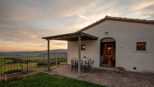 Un petit bâtiment blanc doté d’un patio comprenant une table et des chaises, un espace salon couvert et une vue sur la campagne.