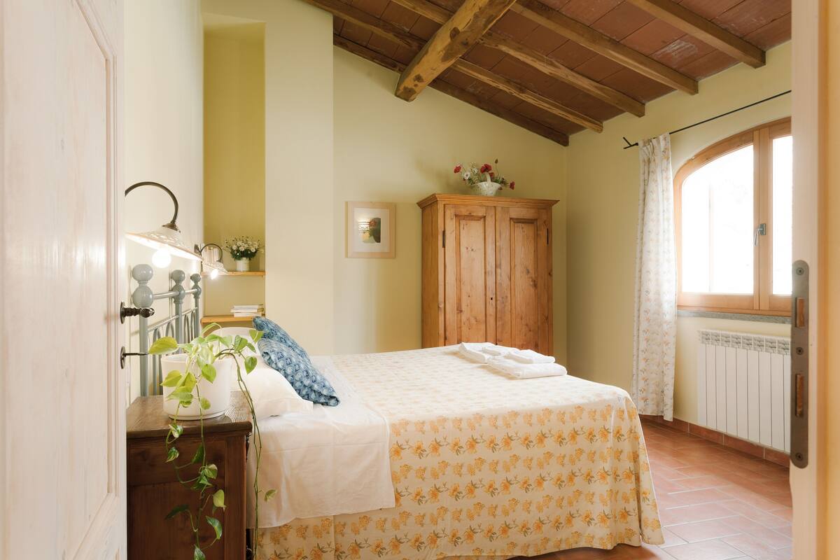 Een slaapkamer met een bed, een houten garderobe, een raam met gordijnen, een nachtkastje met een lamp en een bloempot met een plant.
