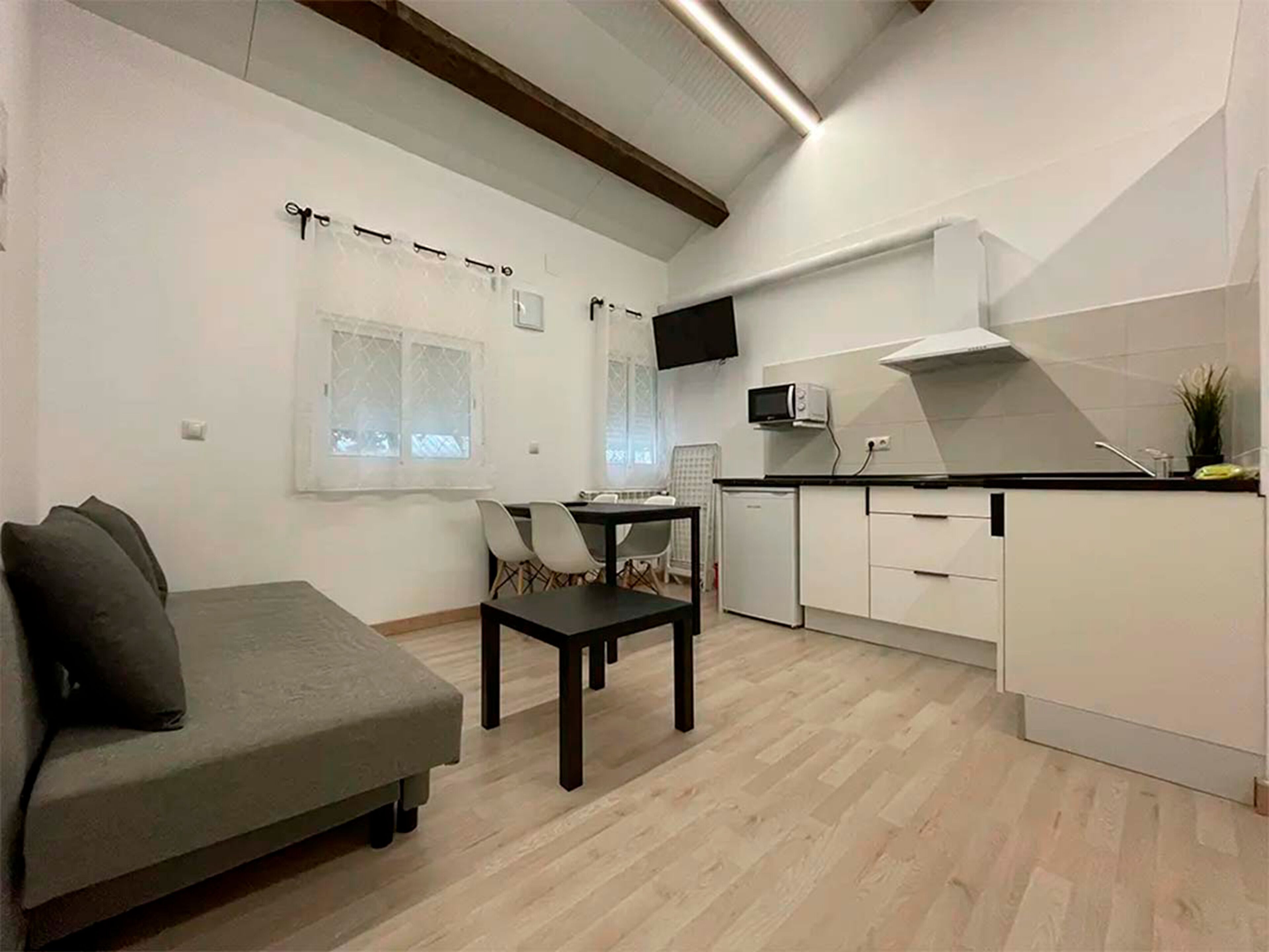 A compact living space with a sofa, dining table, chairs, kitchenette, and a TV mounted on the wall.