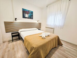 Habitación de hotel con cama, mesita de noche, cabecero, ventana con cortinas, radiador y una obra de arte colgada en la pared.