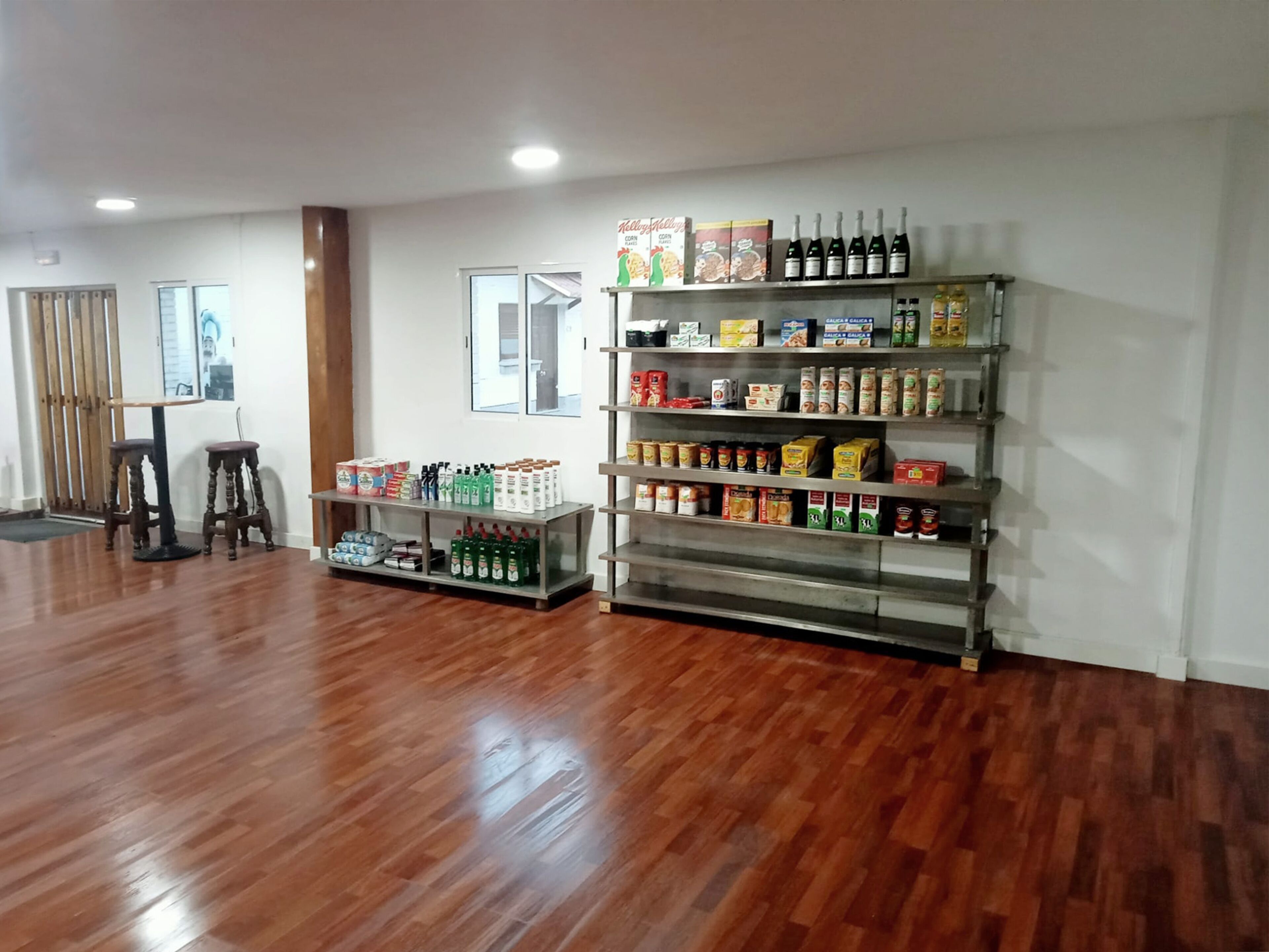 A spacious room with wooden flooring, a bar area with stools, and shelves stocked with beverages and snacks.