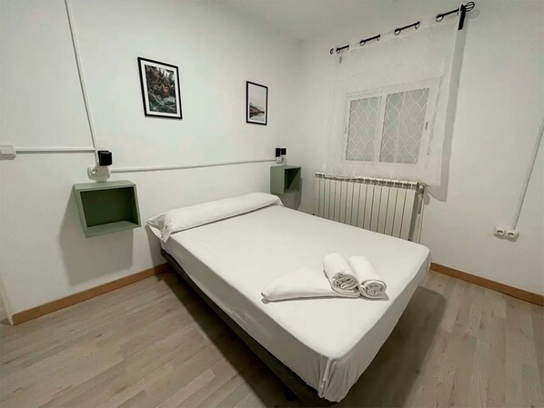 Una cama bien hecha con un colchón y almohadas blancos, una mesita de noche con una lámpara, un cuadro en la pared y una ventana con cortina.