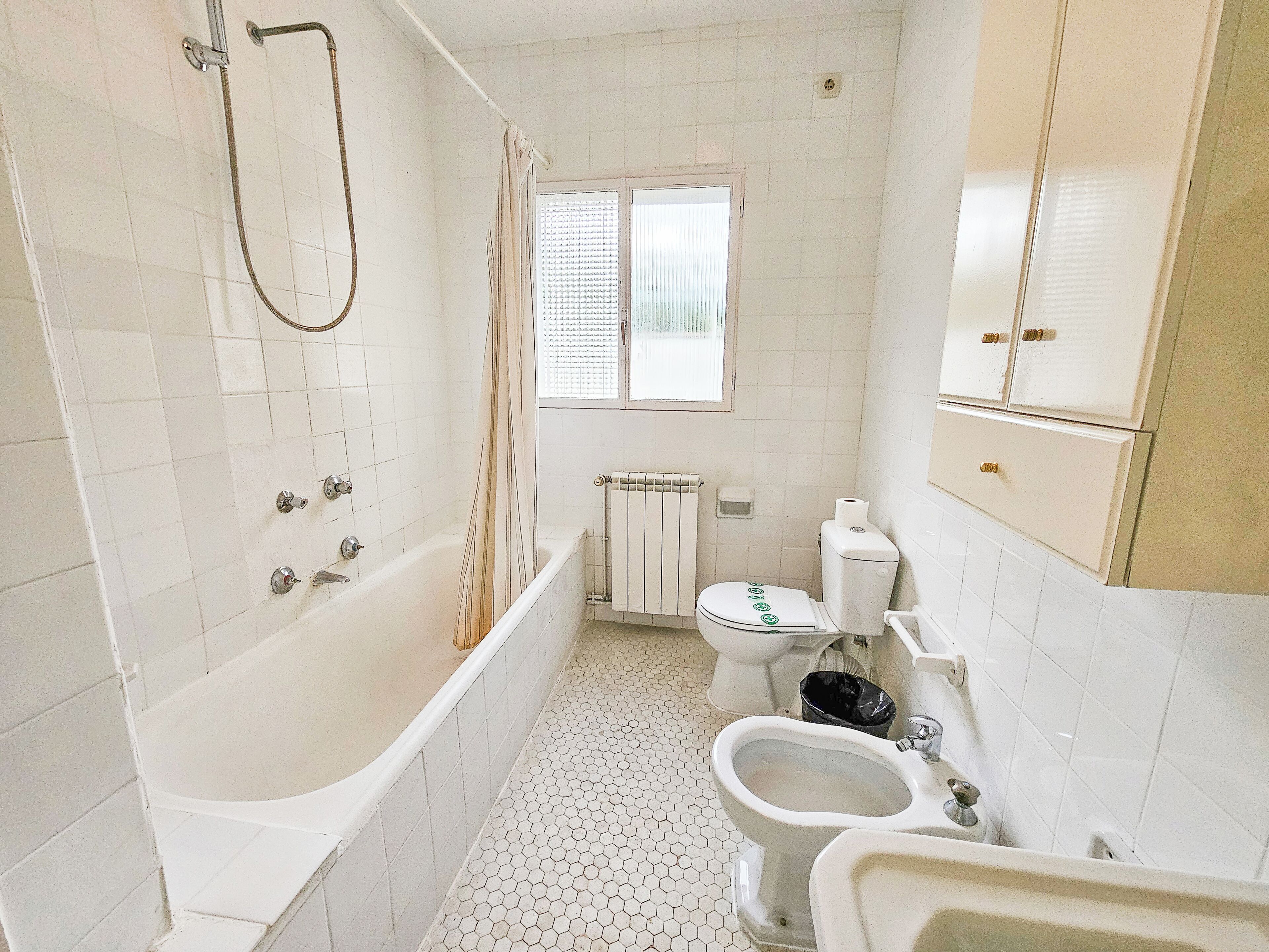 A compact bathroom with a bathtub, toilet, bidet, and a window with blinds.