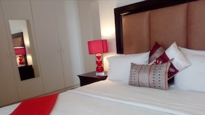 Ein ordentlich bezogenes Bett mit weißen Leinen und dekorativen Kissen, eine rote Tagesdecke sowie ein Nachttisch mit roter Lampe.