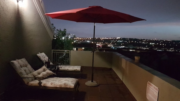 Ein Balkon mit zwei Liegestühlen und einem roten Sonnenschirm, mit Blick auf eine nächtliche Skyline.