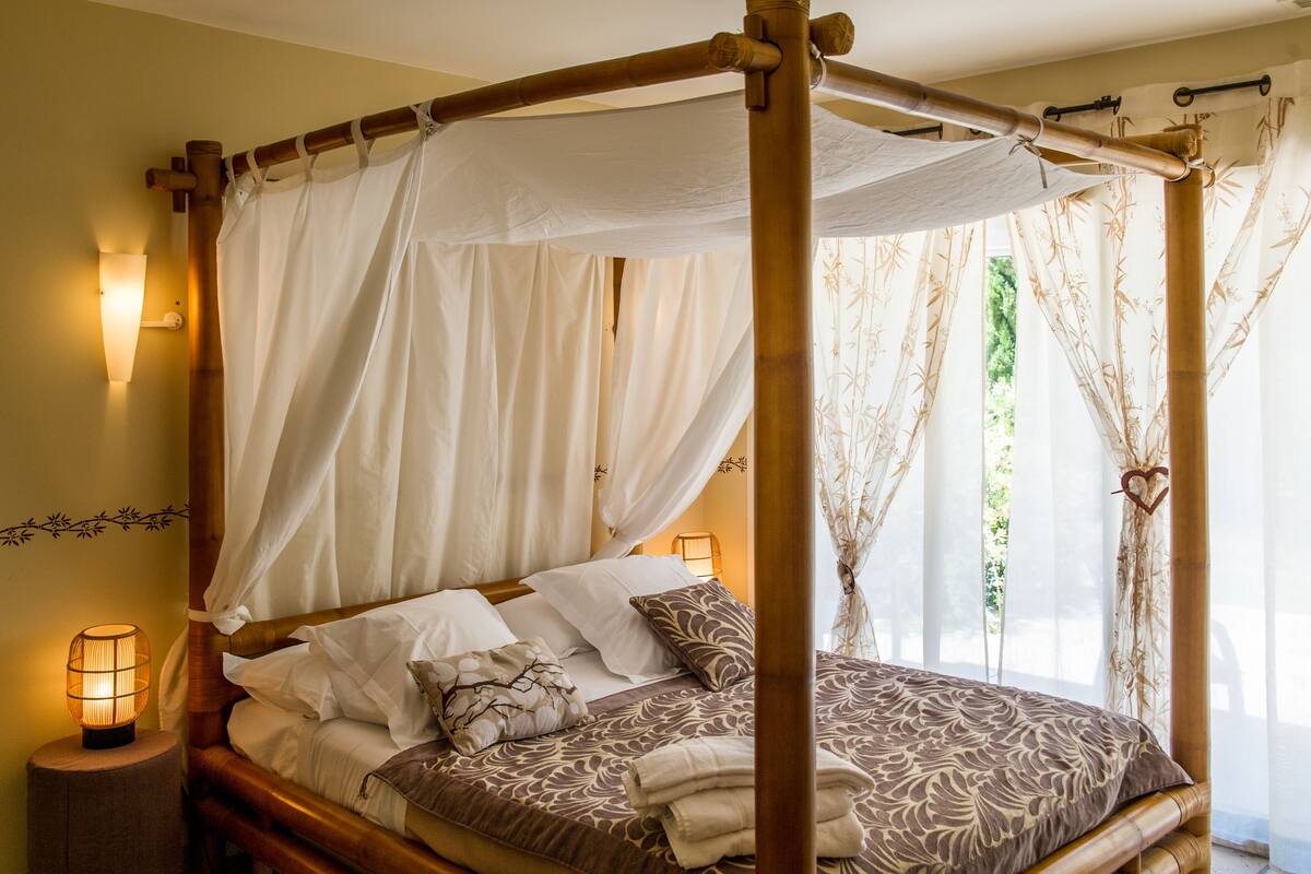 A four-poster bed with white and patterned bedding, a bedside lamp, and a view of greenery through sheer curtains.