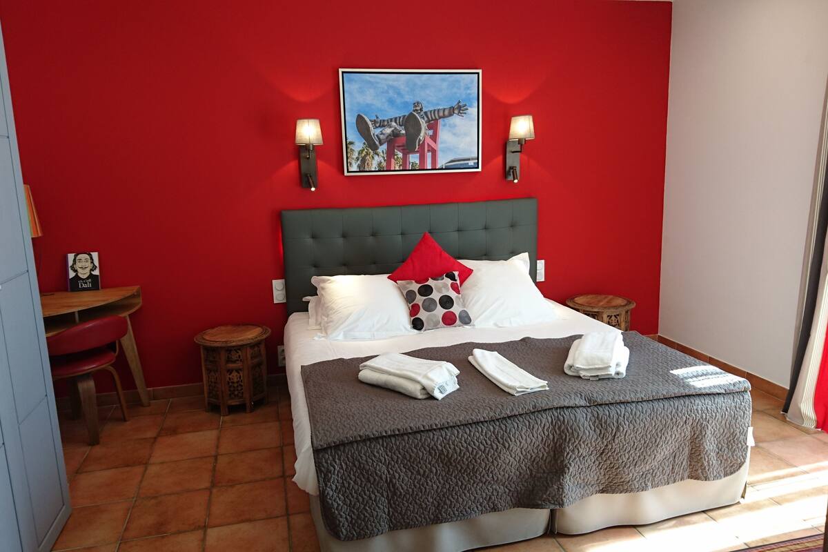 A hotel room with a red wall, a bed with a grey quilt, white pillows, and a painting above the bed. There are two bedside tables with lamps.