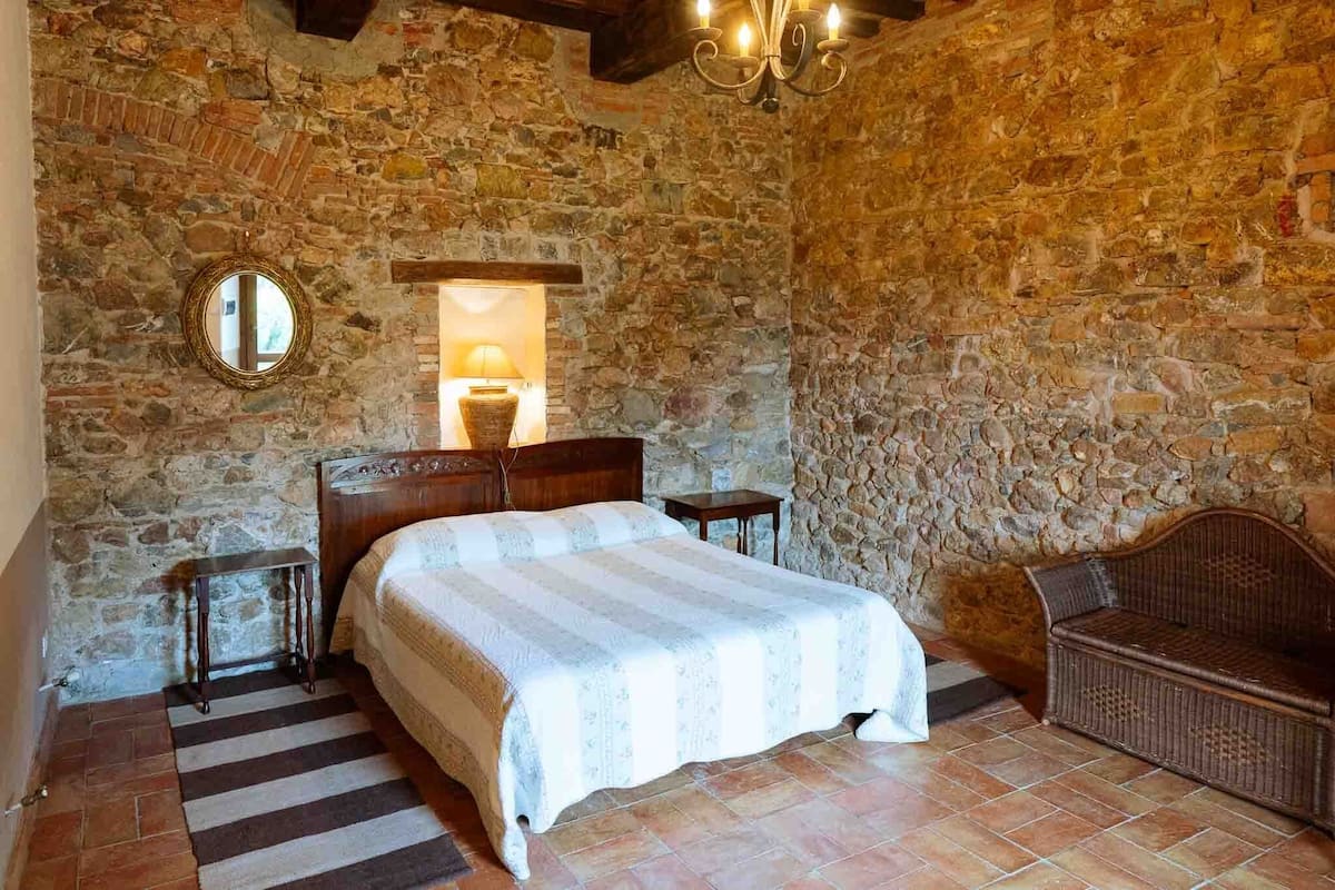 Un dormitorio con paredes de piedra, una cama, una mesita de noche, una silla de mimbre y un espejo.