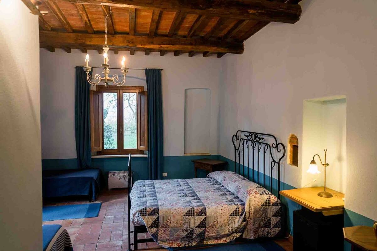 Un dormitorio con cama, techo de madera, ventana con cortinas, mesita de noche con lámpara y un banco.