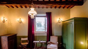 Una habitación con techo de madera, ventana con cortinas rojas, un aparador verde, dos sillas, una mesa pequeña y apliques de pared.