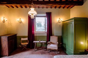 A room with a wooden ceiling, a window with red curtains, a green cabinet, two chairs, a small table, and wall sconces.