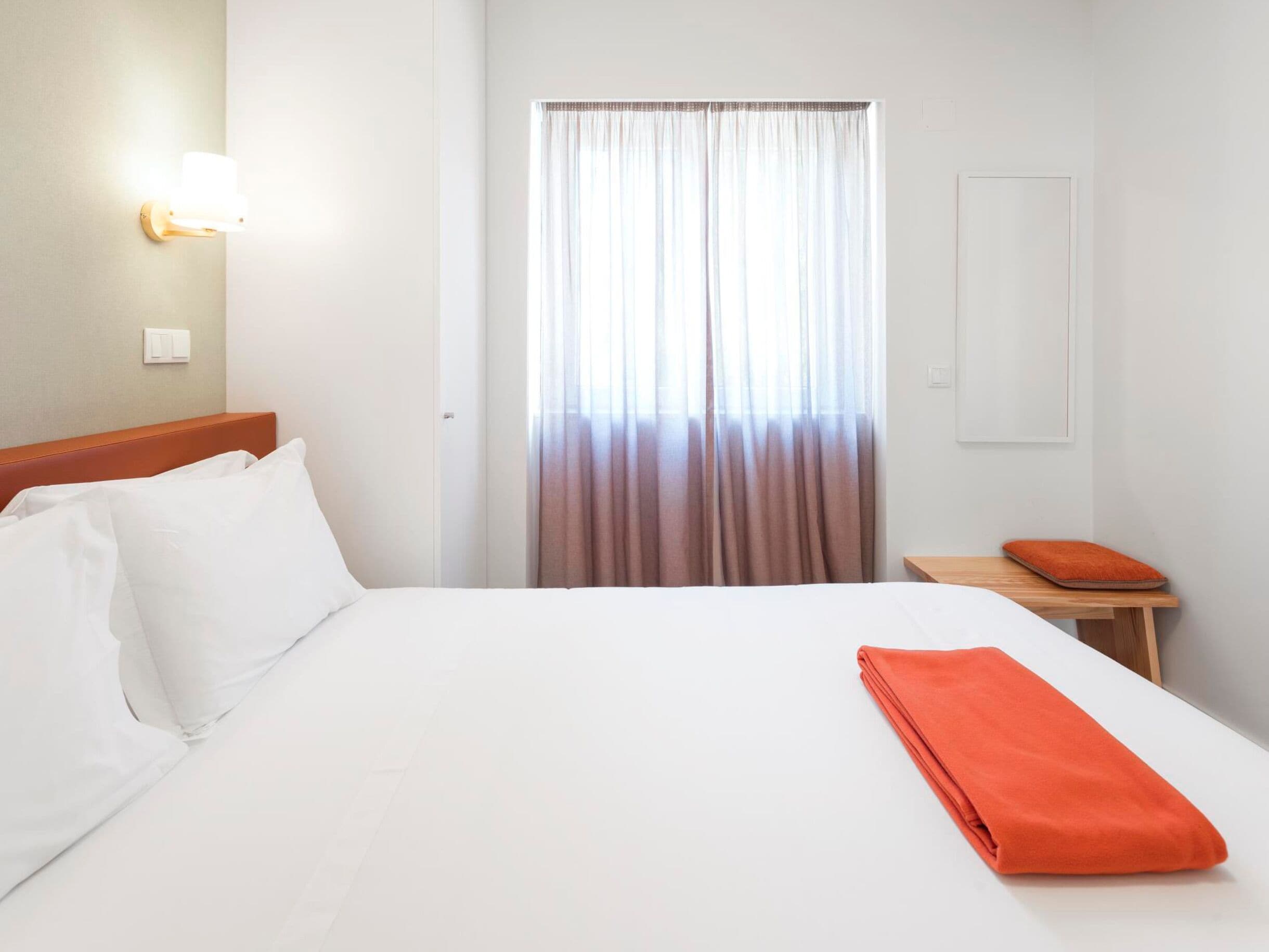 A hotel room with a large bed, a nightstand, a lamp, a window with curtains, and a wall-mounted light.
