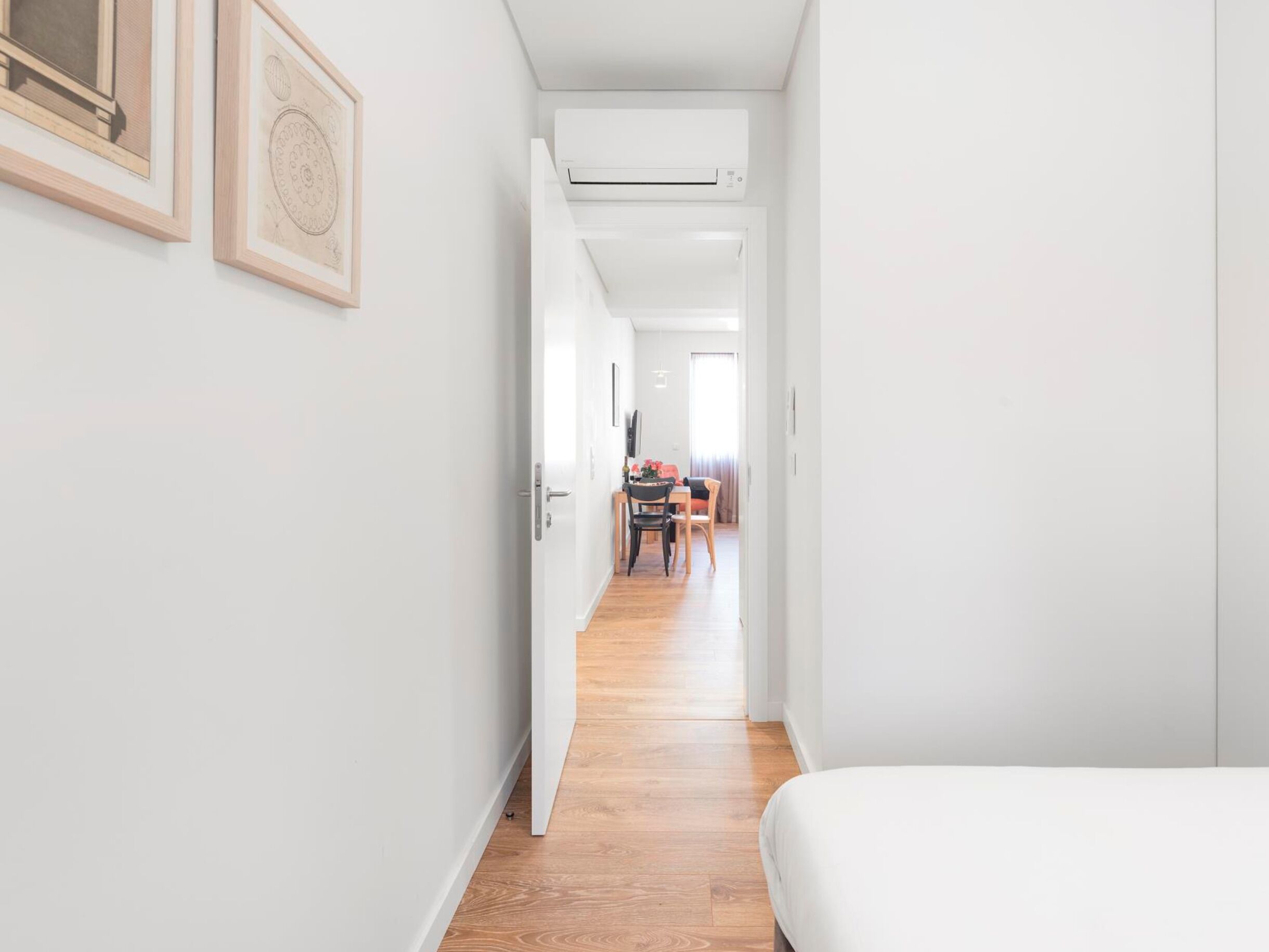A narrow hallway with a wooden floor, white walls, and a bed on the right.