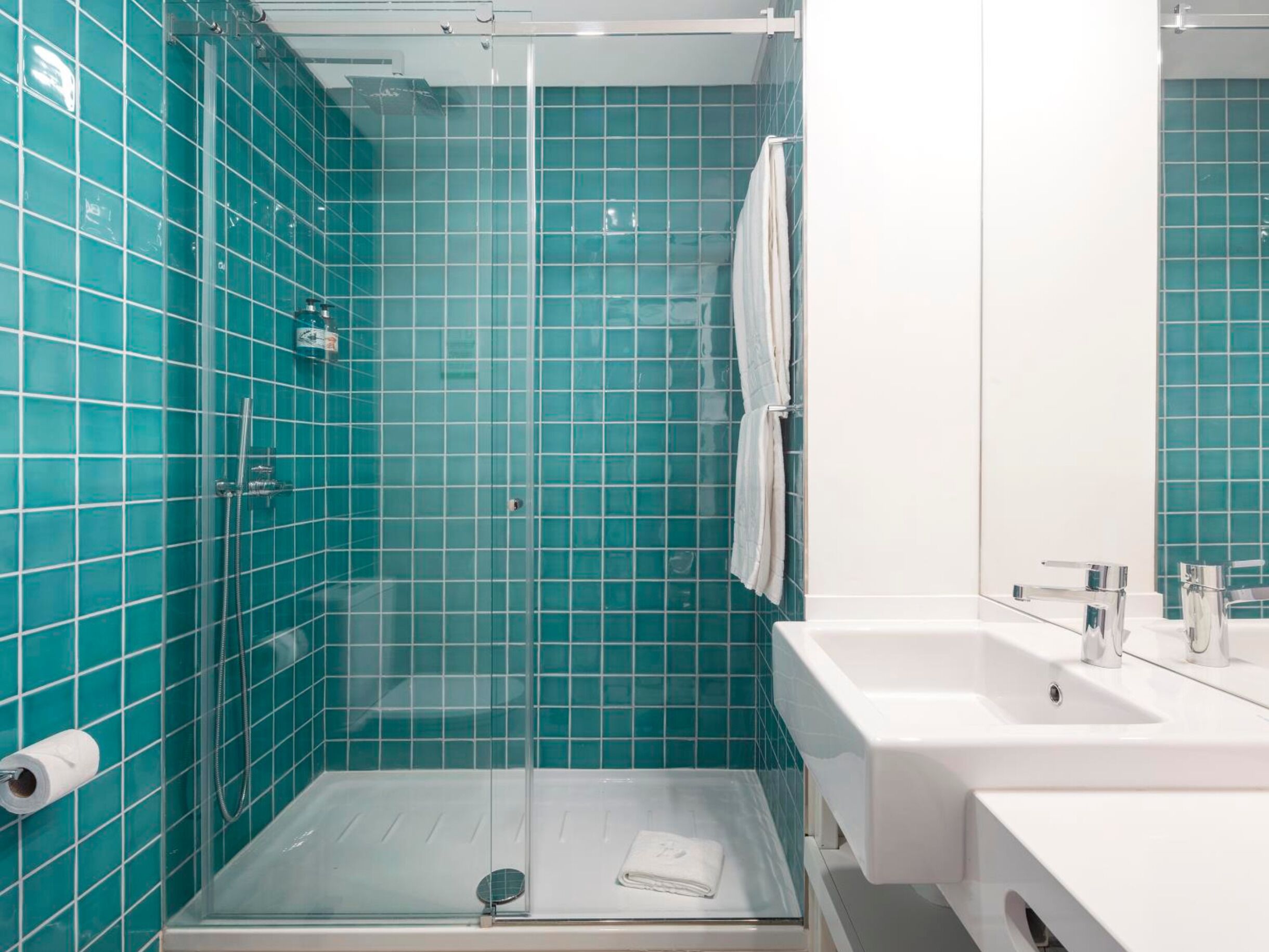 Une salle de bain avec des carreaux de couleur turquoise, une douche en verre, un lavabo blanc et un miroir.