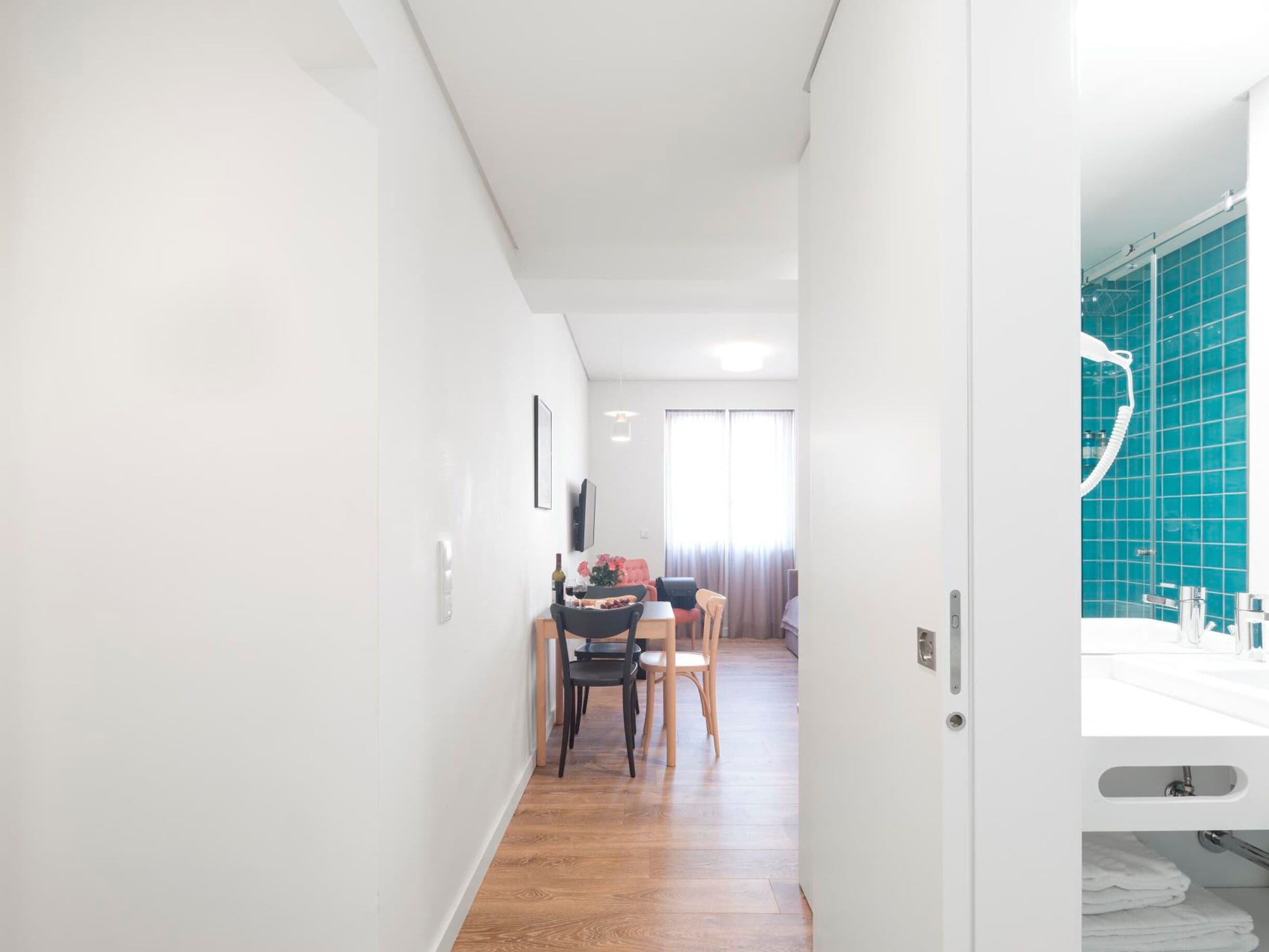 Un couloir mène à une salle à manger équipée d’une table et de chaises, ainsi qu’à une salle de bain avec un lavabo et des murs carrelés.