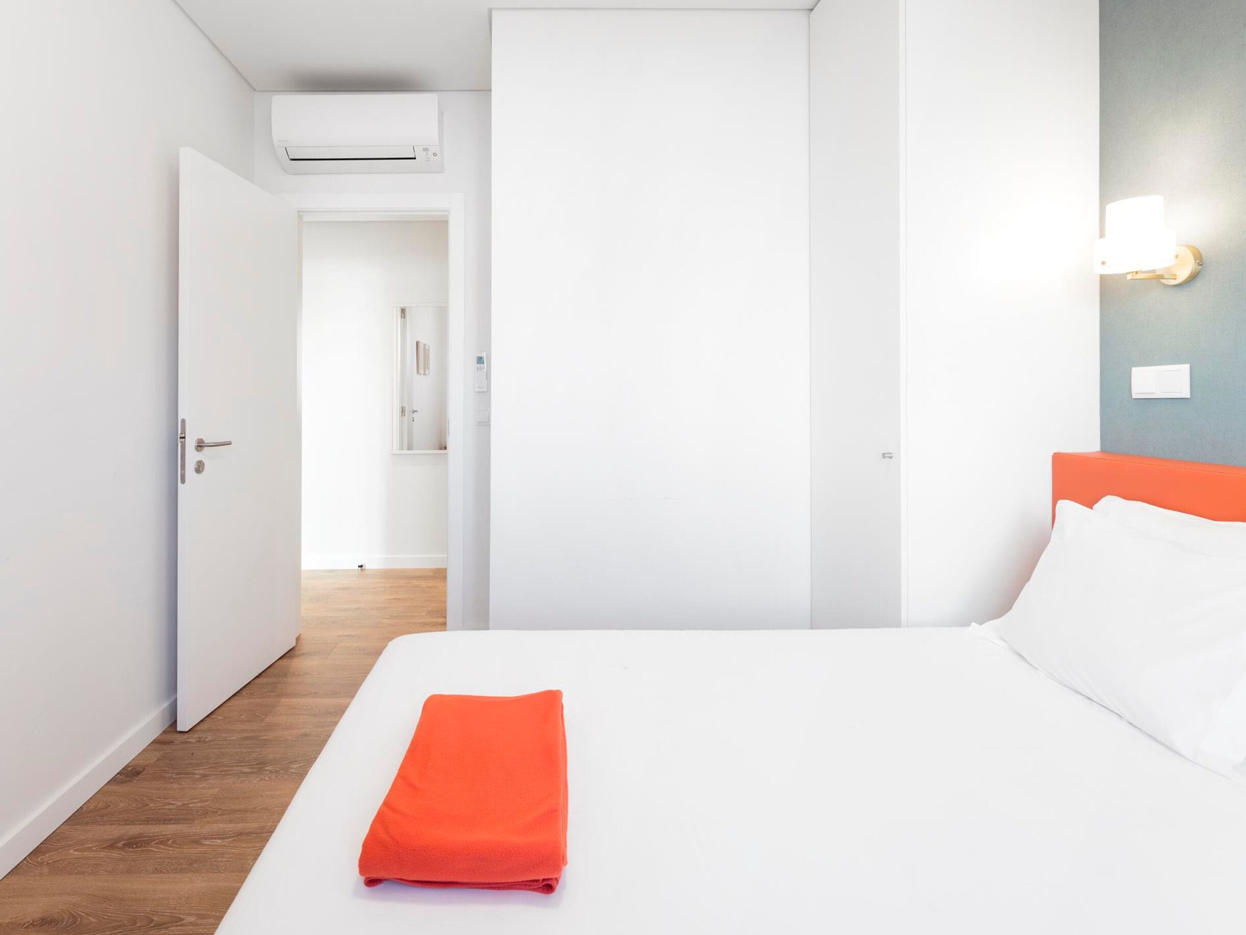 Hotelkamer met een bed, een feloranje handdoek, wit beddengoed, een houten vloer, een witte deur, een witte muur, een lamp en een airconditioner.
