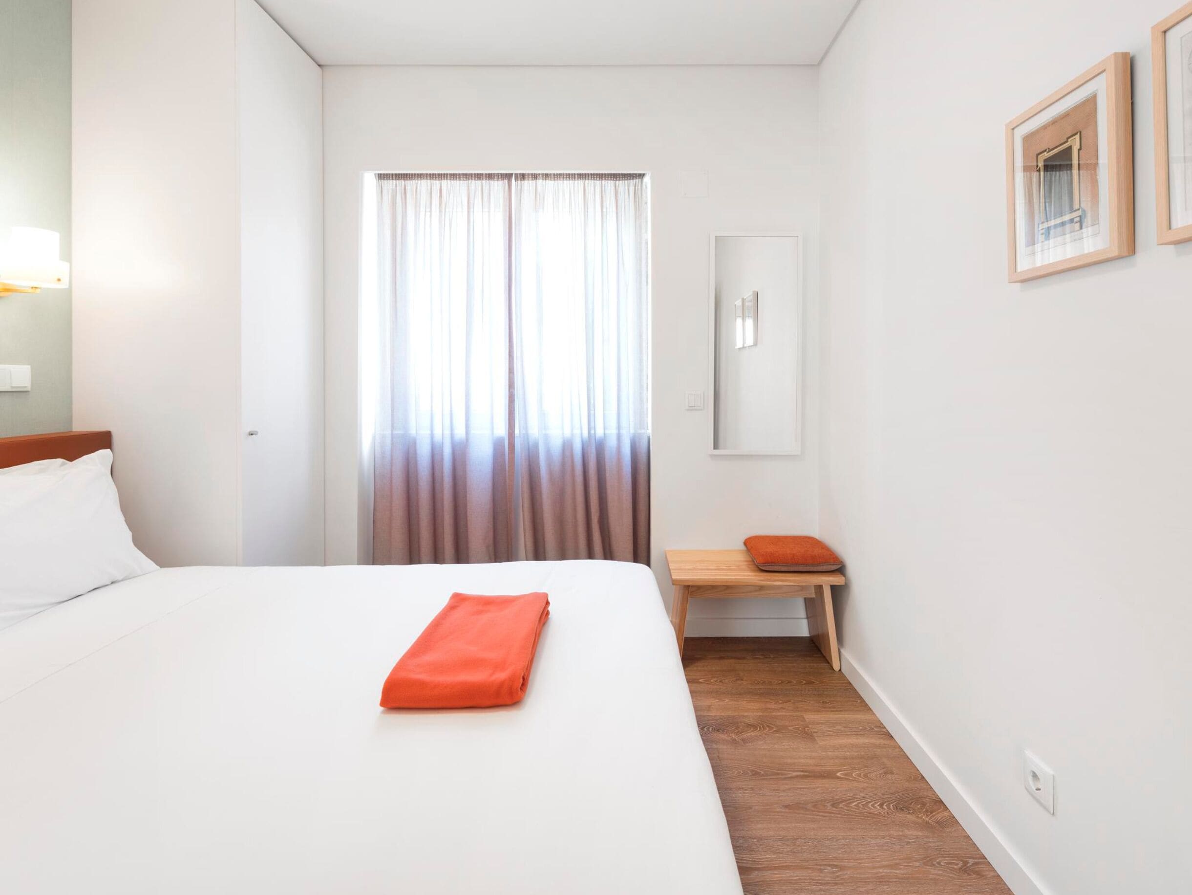 A hotel room with a bed, a wooden nightstand, a mirror, and a window with curtains.