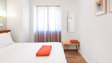A hotel room with a bed, a wooden nightstand, a mirror, and a window with curtains.