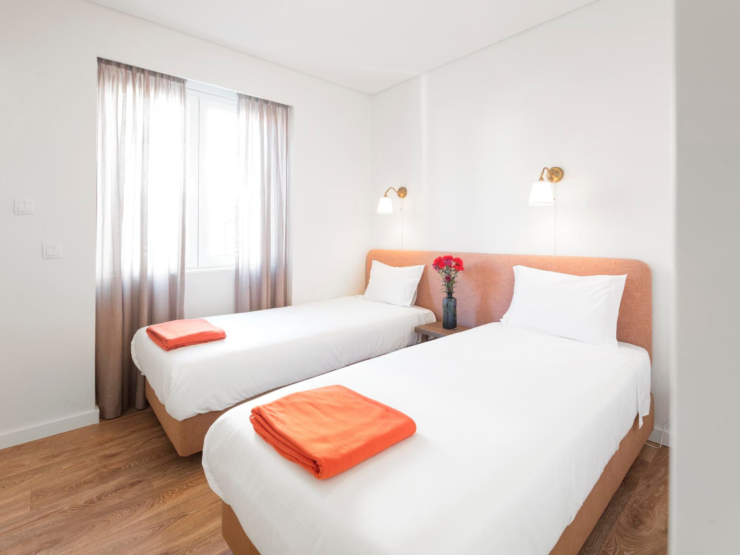 Una habitación de hotel con dos camas, una ventana con cortinas y un florero con flores en la mesita de noche.