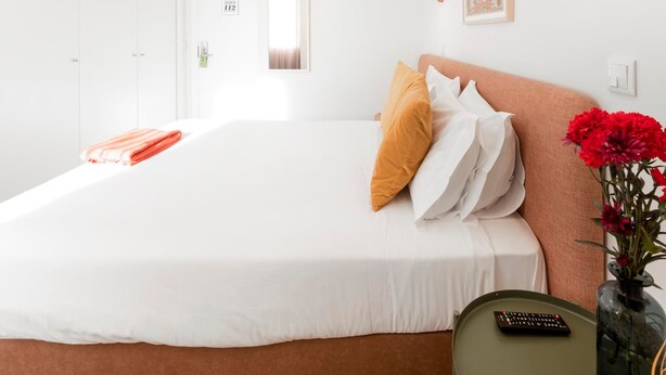 A hotel room with a large bed, a bedside table with a remote control, a vase with flowers, and a picture on the wall.