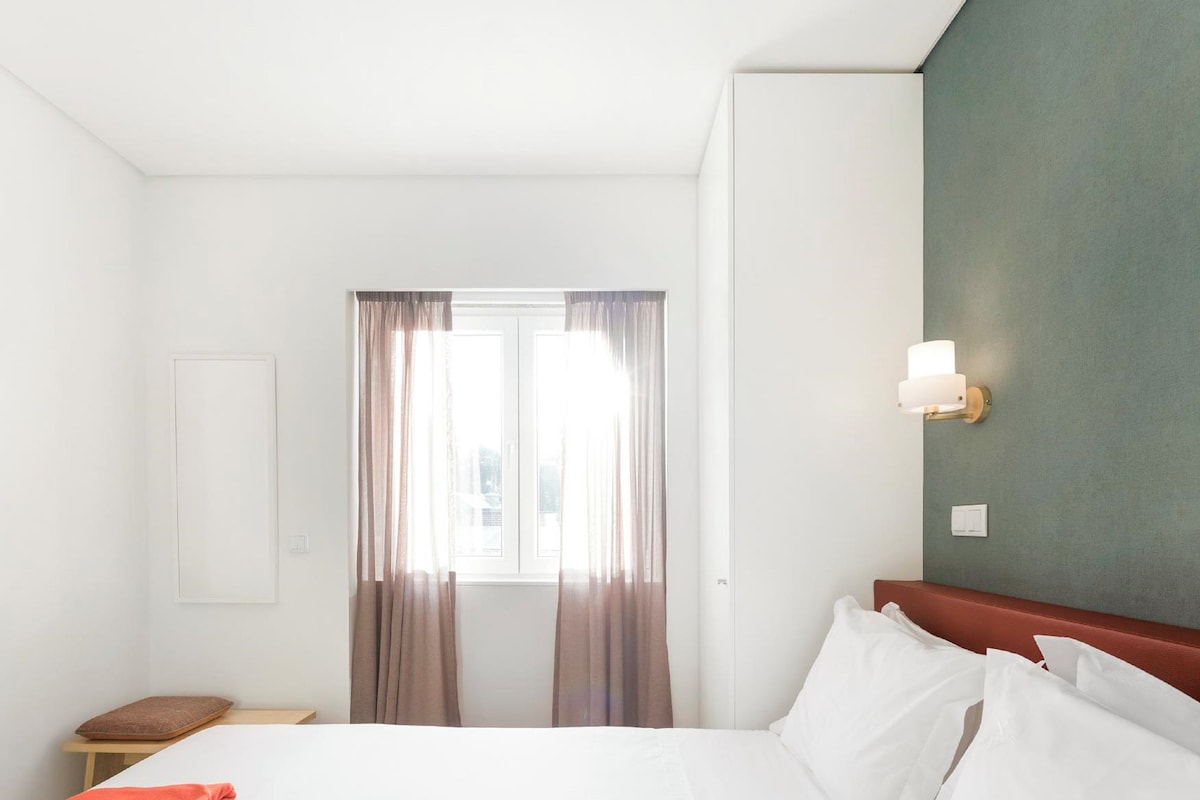 Una habitación de hotel con una cama grande, una mesita de noche, una lámpara de pared y una ventana con cortinas.