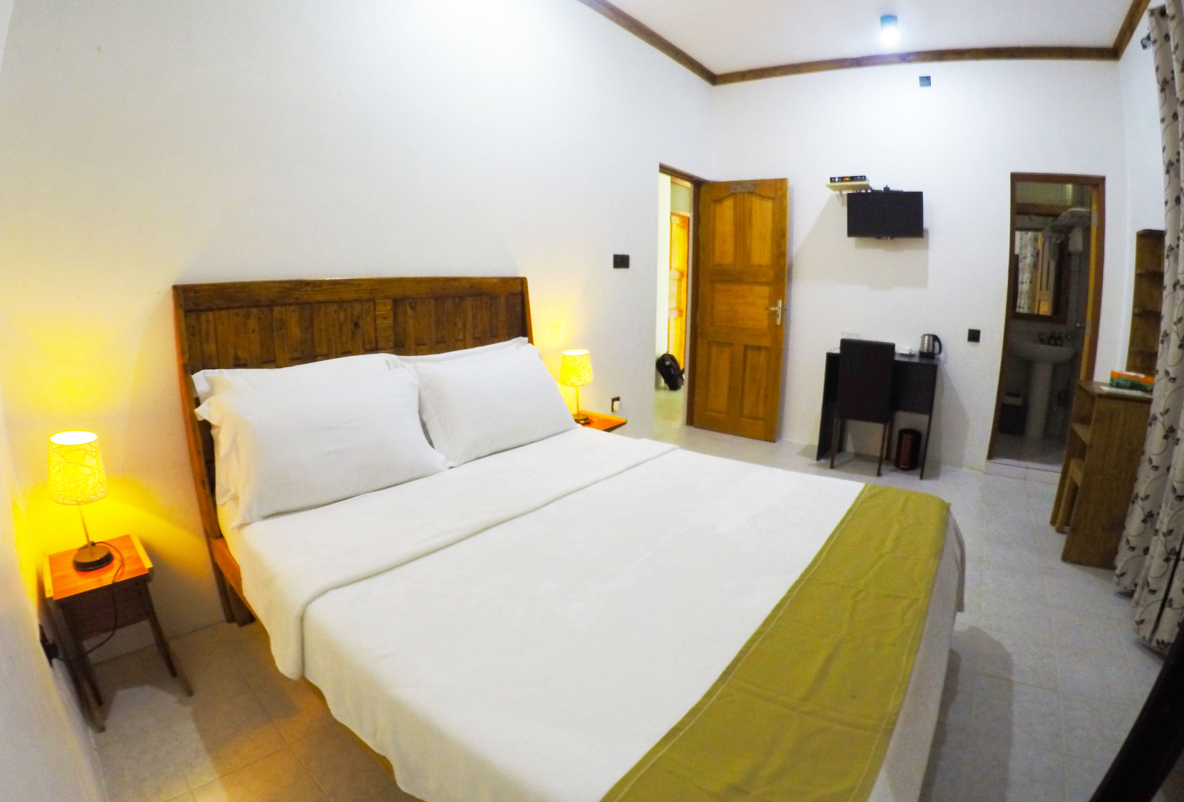 A hotel room with a large bed, a wooden headboard, a bedside table with a lamp, a small desk with a chair, and a mirror on the wall.