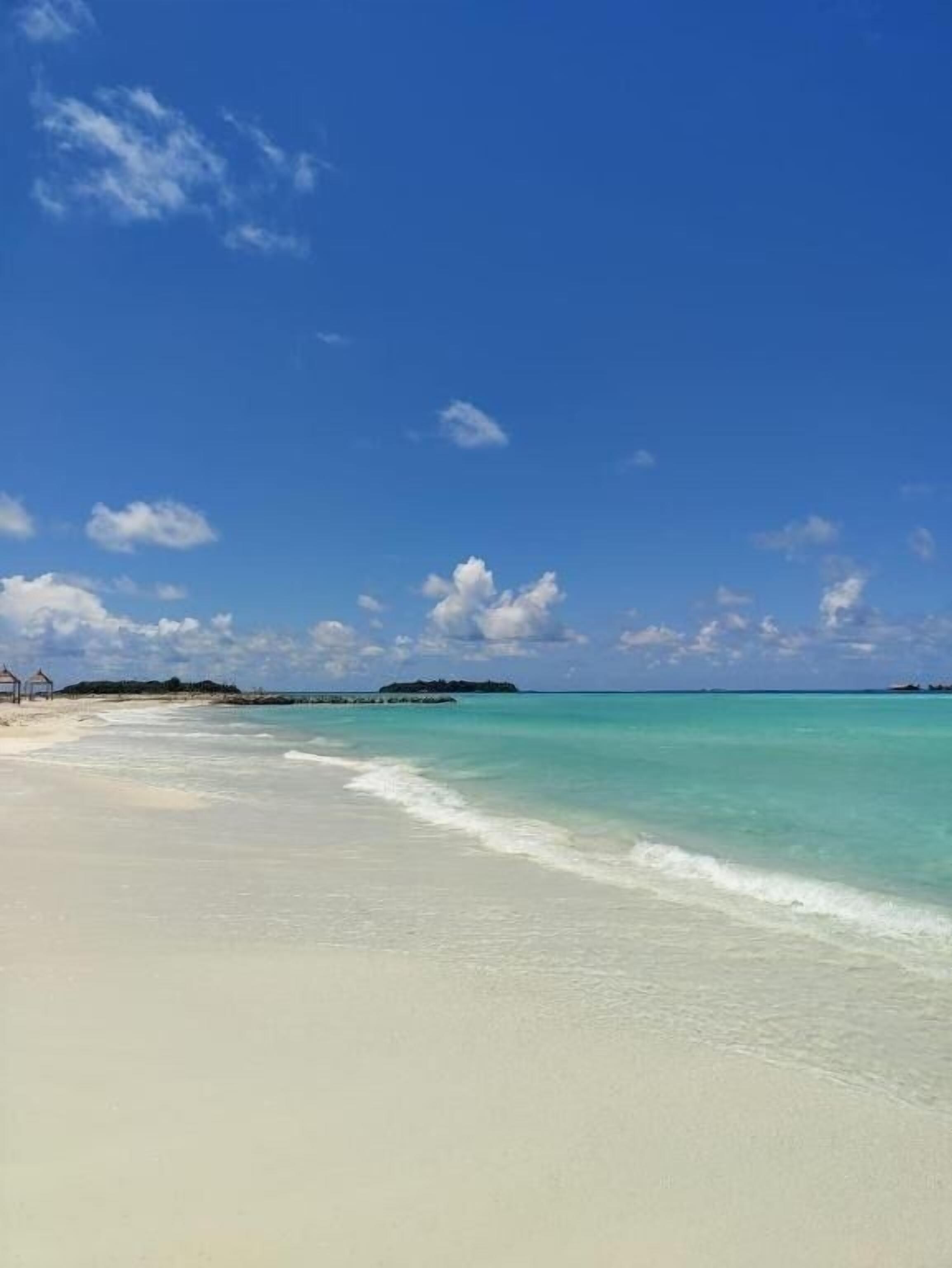 A beach with clear turquoise water, a sandy shore, and a few structures in the distance.