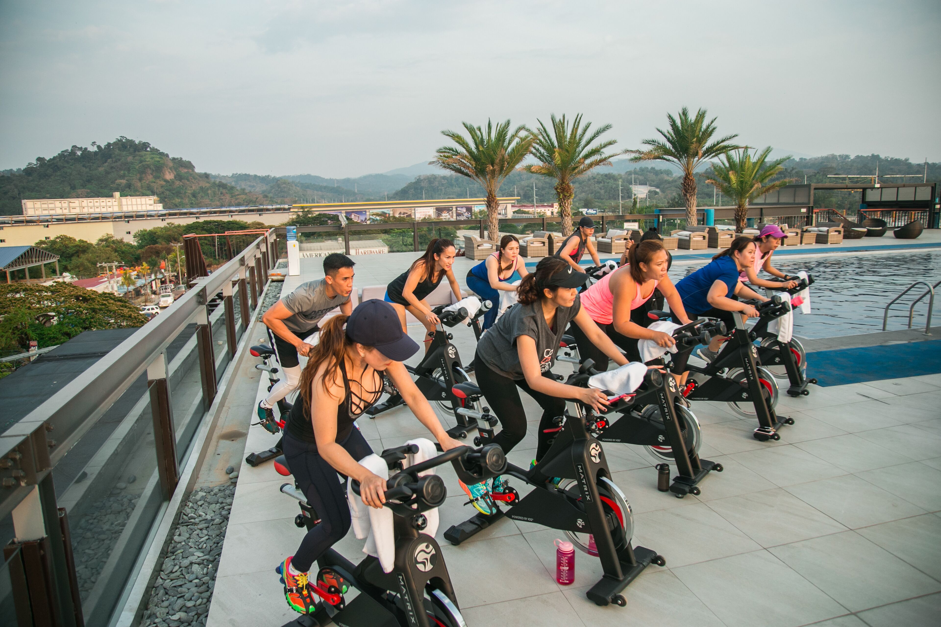 A group of people exercising on stationary bikes near a pool, with a view of hills and a bridge in the background.