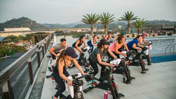 A group of people exercising on stationary bikes near a pool, with a view of hills and a bridge in the background.