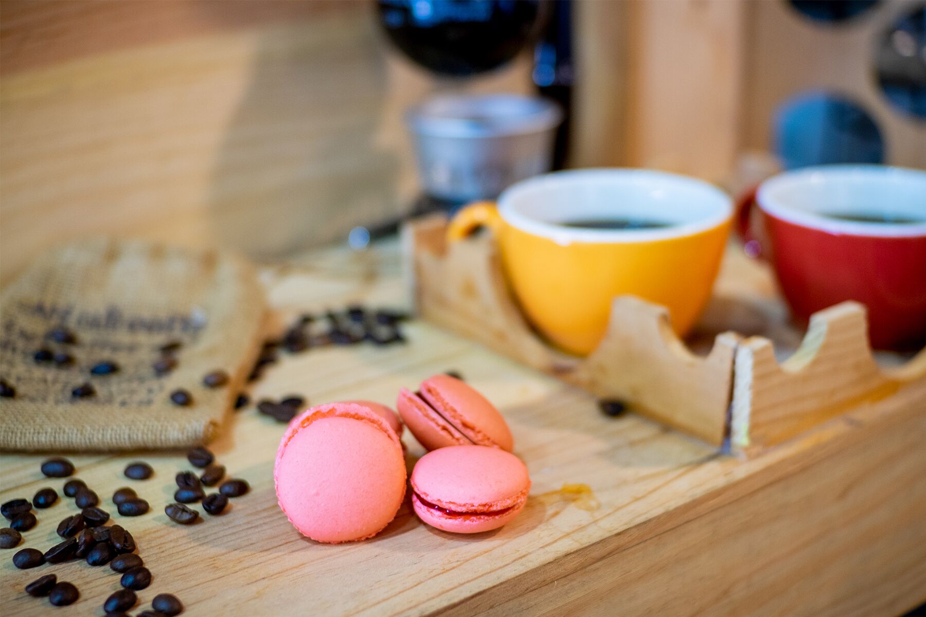 A wooden table with pink macarons, coffee beans, and colorful cups.
