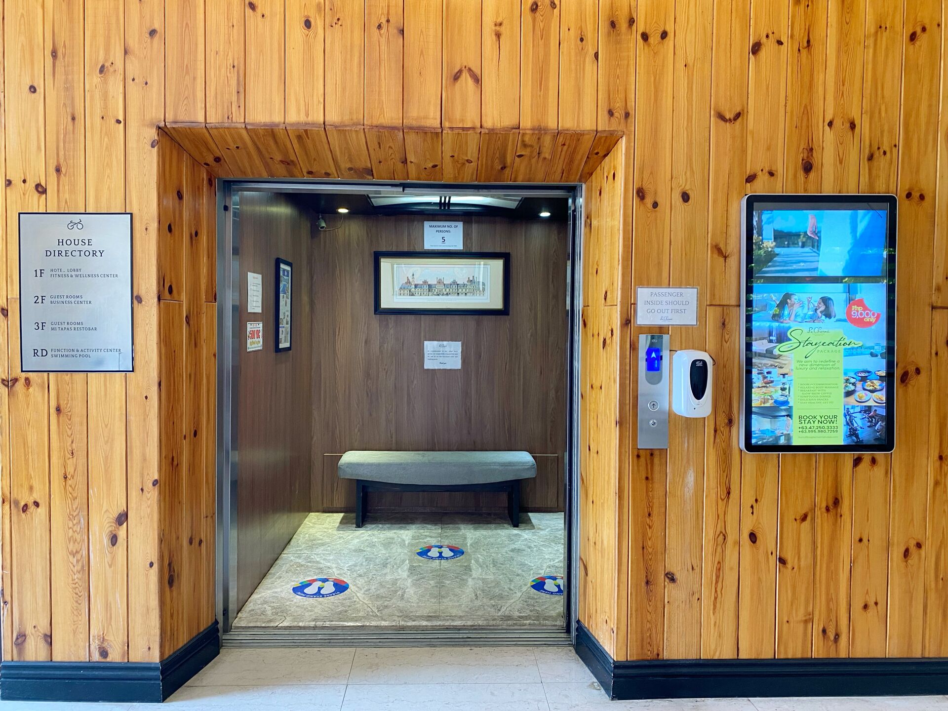 An elevator interior with a wooden wall, a directory sign, a framed picture, a bench, and a digital display.