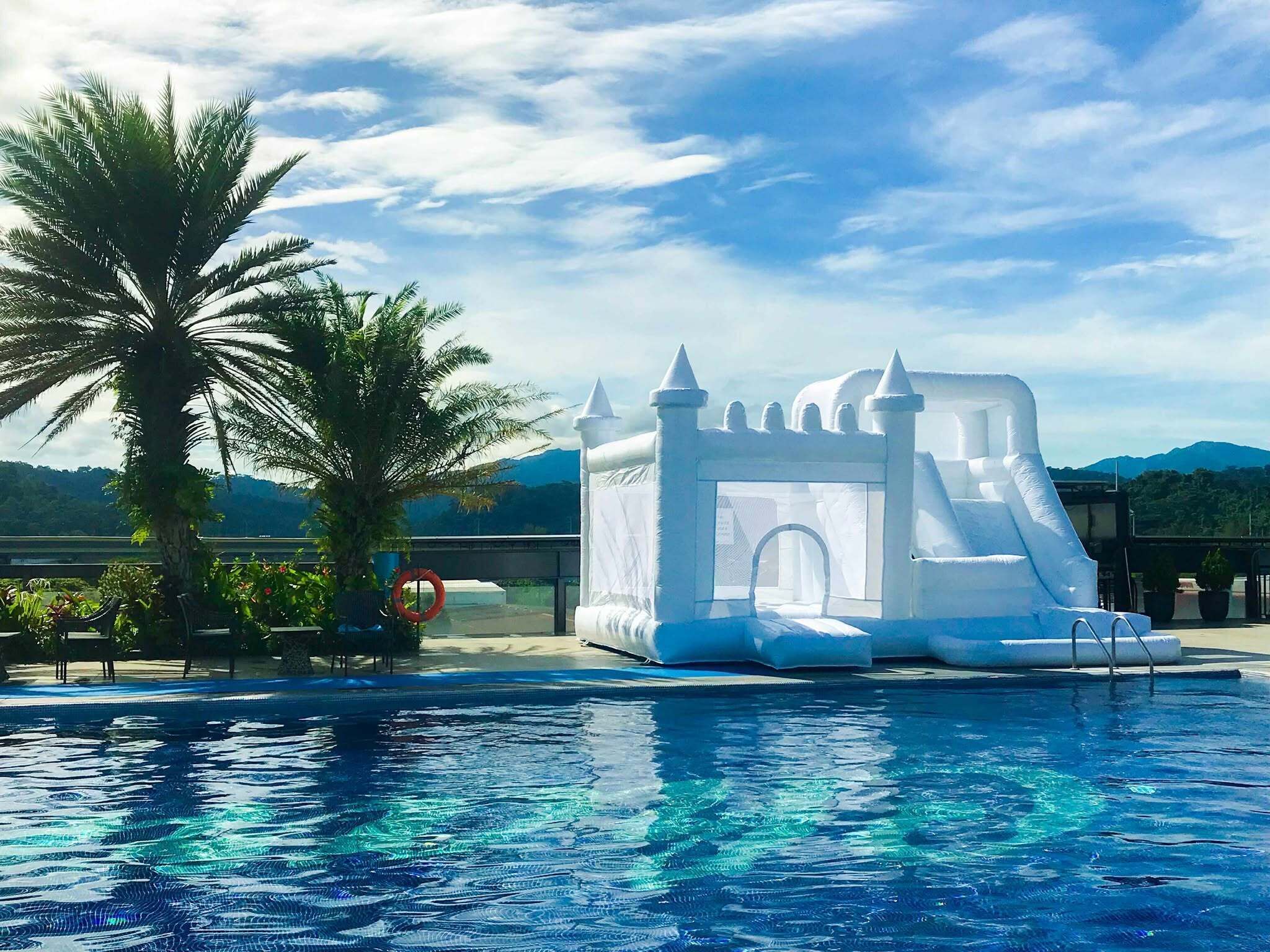 Un castillo inflable junto a una piscina con palmeras y vista a la montaña.