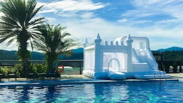 Un castillo inflable junto a una piscina con palmeras y vista a la montaña.