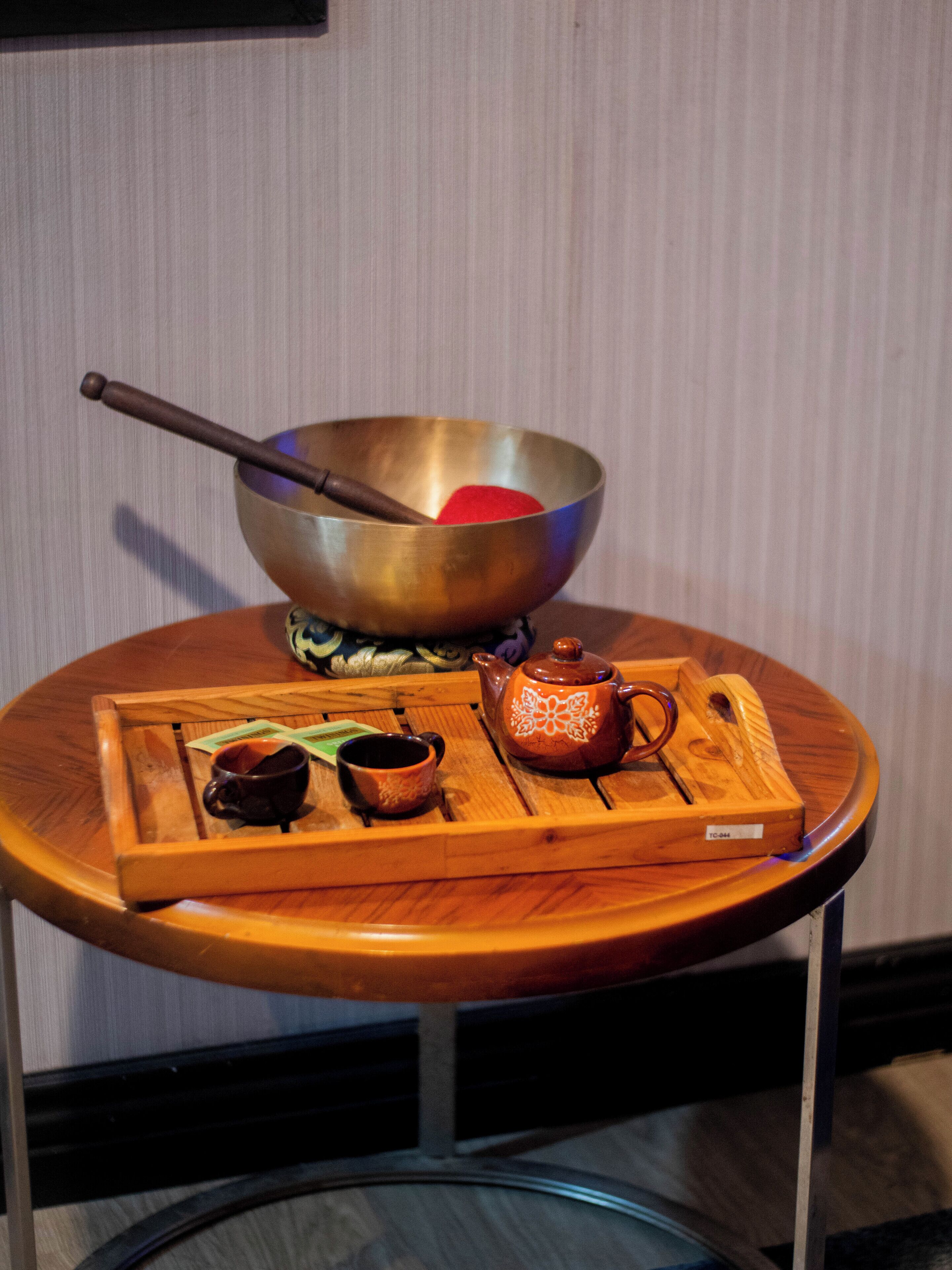A round wooden table with a metal bowl, a wooden spoon, a teapot, and two cups.