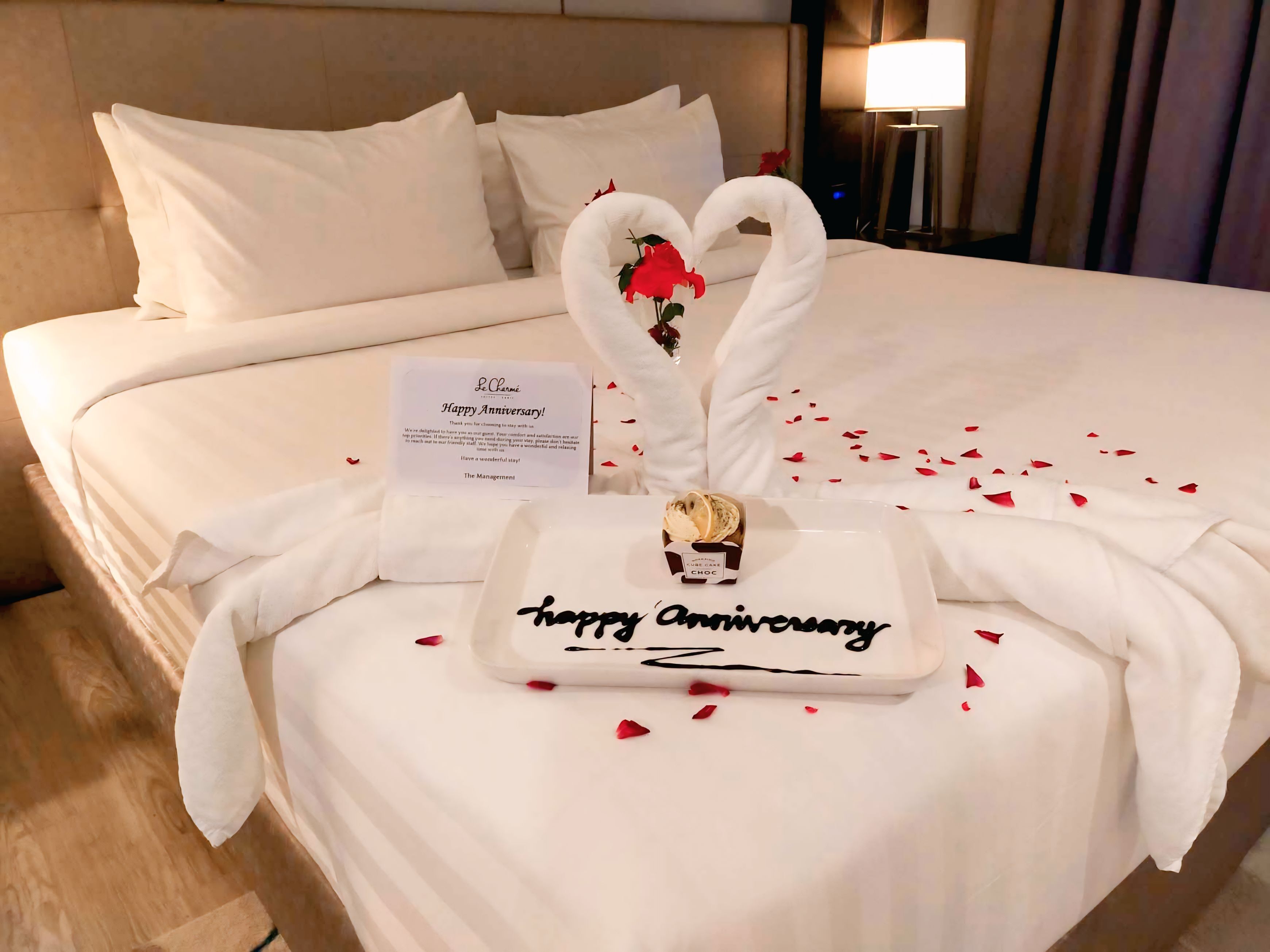 A hotel room with a neatly made bed, a sign reading "Happy Anniversary," and a chocolate cake on the bed.