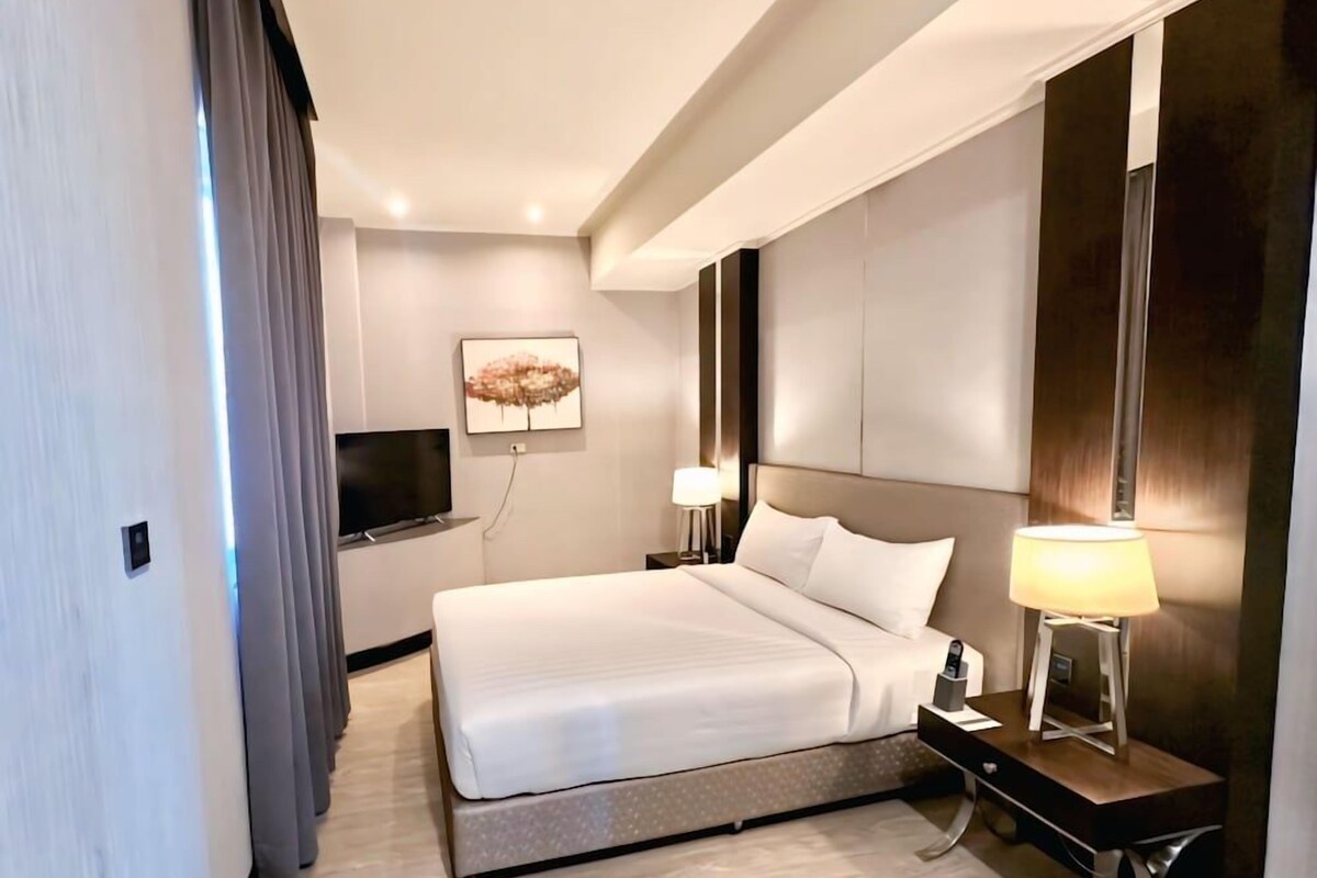 Una habitación de hotel moderna con una cama grande, un televisor de pantalla plana, una mesita de noche con lámpara y una obra de arte colgada en la pared.